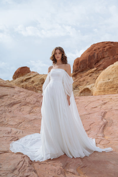 A woman in a flowing white Noa - Jenny Yoo Wedding Dress gown with dramatic sleeves stands on a rocky desert terrain under a cloudy sky.
