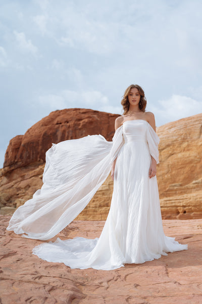 Woman in a flowing Jenny Yoo Wedding Dress gown with dramatic sleeves, standing against a desert rock formation backdrop.
