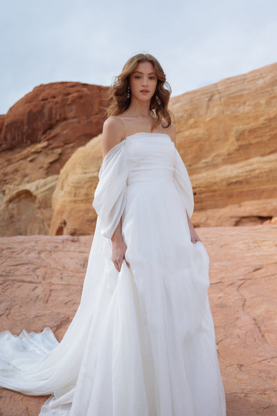 A woman in a white Jenny Yoo Wedding Dress gown with dramatic sleeves standing against a desert rock formation.