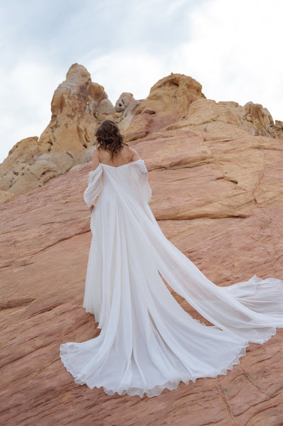 A woman in an elegant Jenny Yoo Wedding Dress with dramatic sleeves and a flowing train stands on rocky terrain, looking away from the camera.