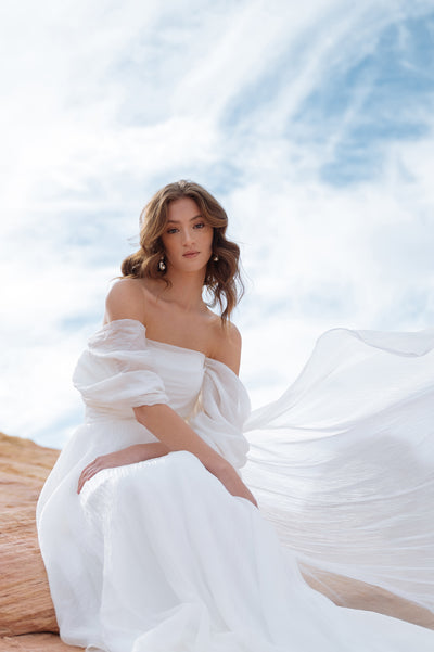 A woman in a flowing Jenny Yoo Wedding Dress gown seated on a sandy terrain with a cloudy sky in the background.