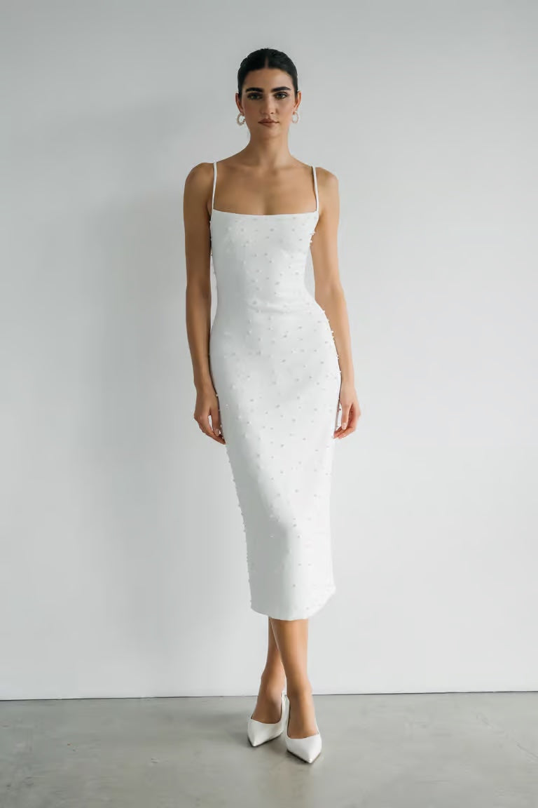 A person poses against a white wall in the Bergamot Bridal Oksana - Jenny Yoo Little White Dress, a fitted luxe stretch crepe midi with thin straps and sleeveless design, styled with white pointed-toe heels.