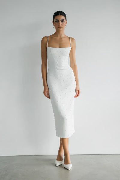 A person poses against a white wall in the Bergamot Bridal Oksana - Jenny Yoo Little White Dress, a fitted luxe stretch crepe midi with thin straps and sleeveless design, styled with white pointed-toe heels.
