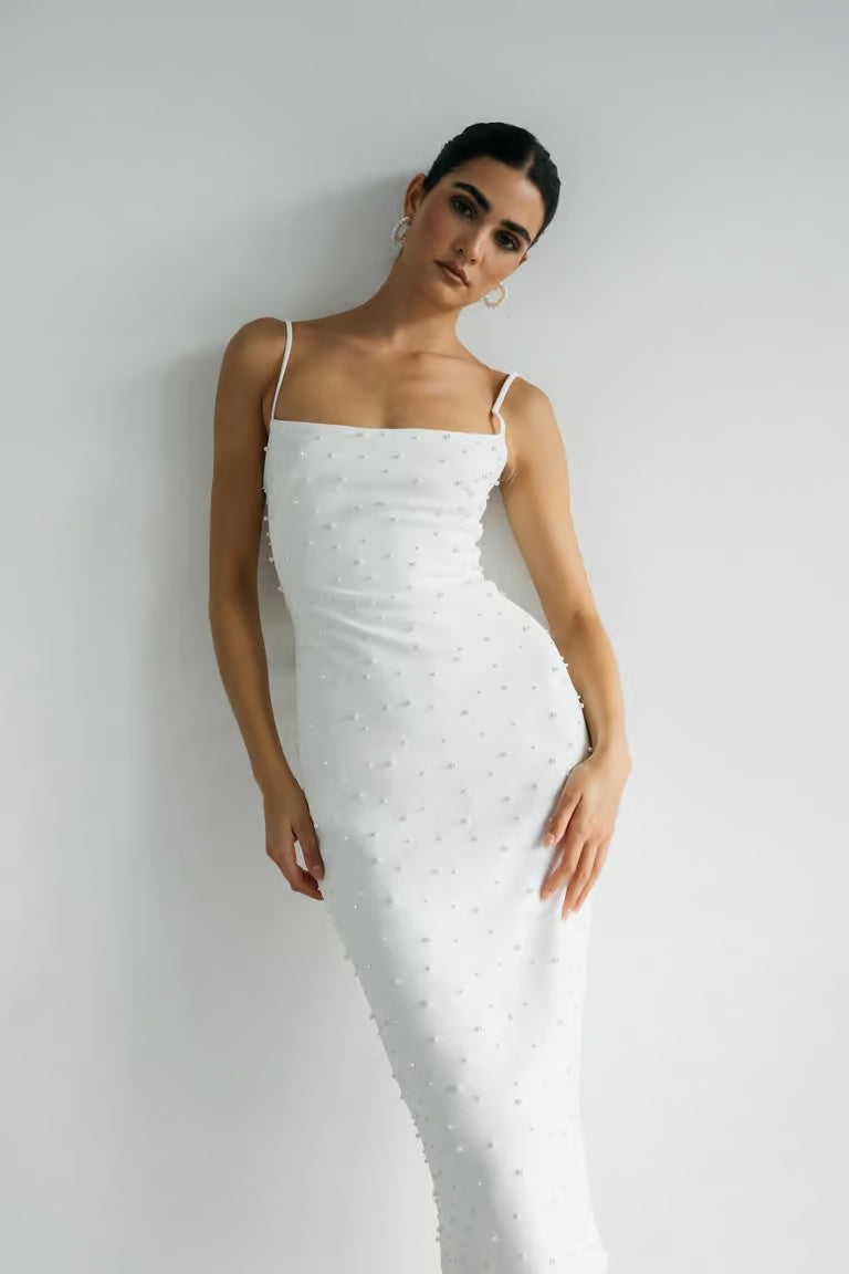 A woman stands against a plain white wall wearing the Oksana - Jenny Yoo Little White Dress by Bergamot Bridal—a fitted, sleeveless white dress in luxe stretch crepe with a delicate pearl scatter.