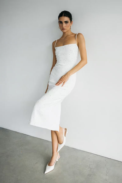 A woman wears the Bergamot Bridal Oksana - Jenny Yoo Little White Dress, a fitted white midi in luxe stretch crepe with thin straps and pearl details, styled with white pointed-toe heels against a plain white wall on concrete.