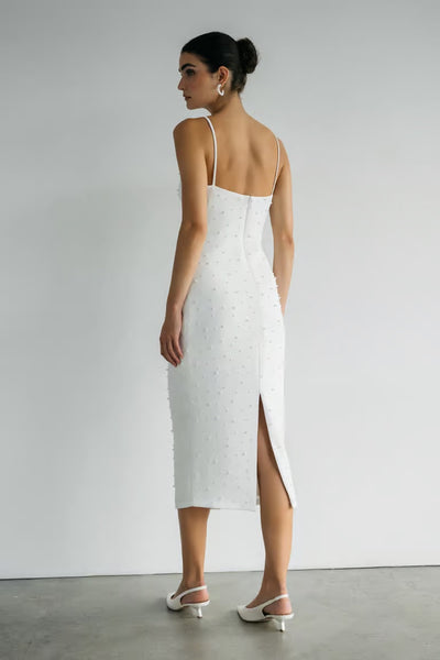 A woman in the Oksana - Jenny Yoo Little White Dress by Bergamot Bridal, crafted from luxe stretch crepe with thin straps and a back slit, stands indoors facing away. She pairs the dress with white heels and wears her hair in a bun.