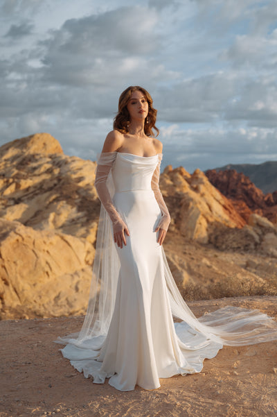 A woman in an elegant Jenny Yoo Wedding Dress made of luxe satin fabric, with an off the shoulder sleeve, stands in a desert landscape at sunset.