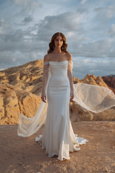 A woman in an elegant off-the-shoulder sleeve gown stands against a rocky desert backdrop, with her veil caught in a gentle breeze. The gown is the Olivia from Bergamot Bridal by Jenny Yoo.