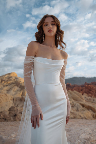 A woman in an elegant white Jenny Yoo Wedding Dress gown made from luxe satin fabric, posing against a rugged outdoor backdrop.
