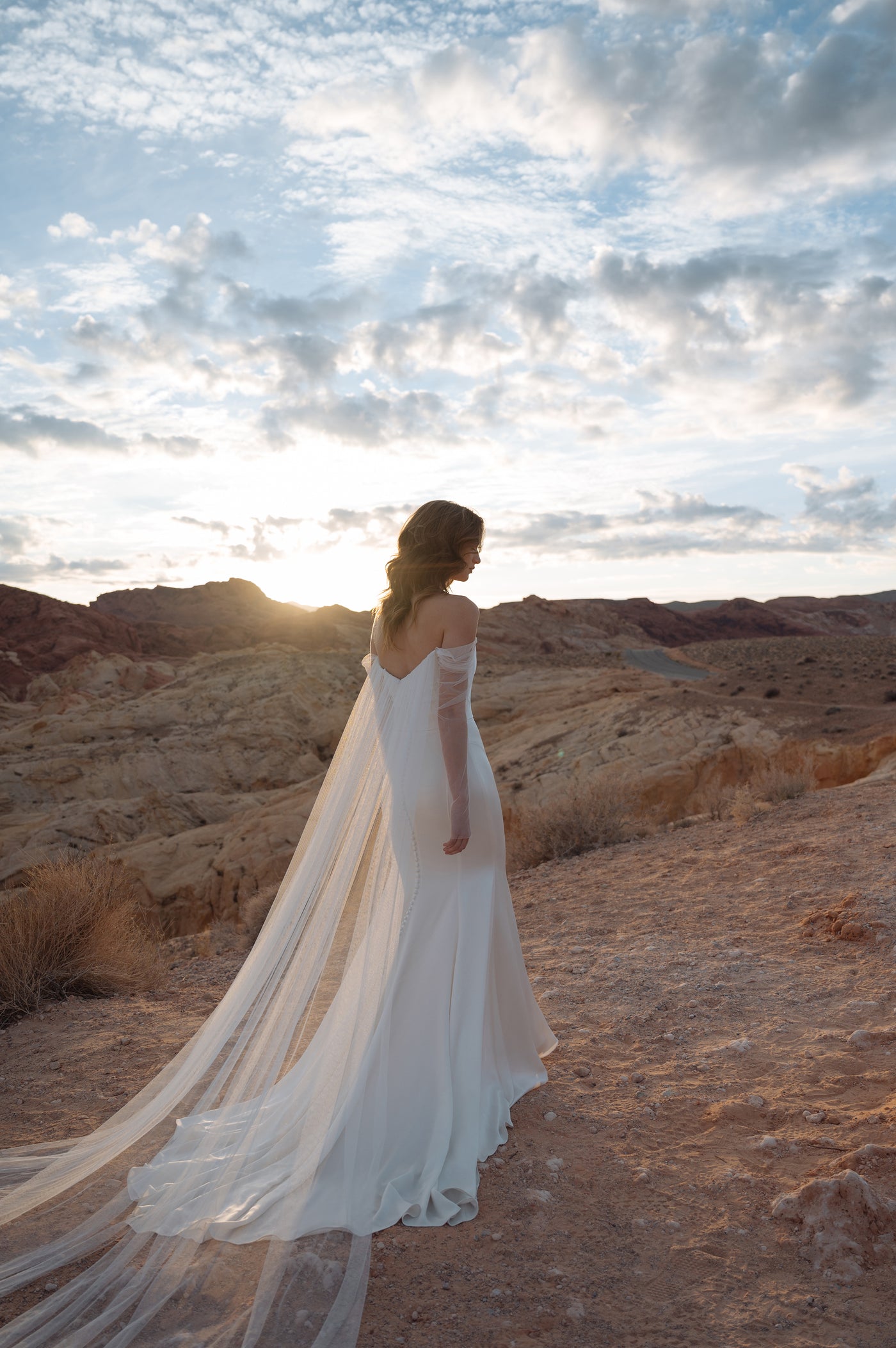 A bride in a Jenny Yoo Wedding Dress gown, featuring a fit and flare skirt and luxe satin fabric, stands contemplatively at a desert location at sunset.