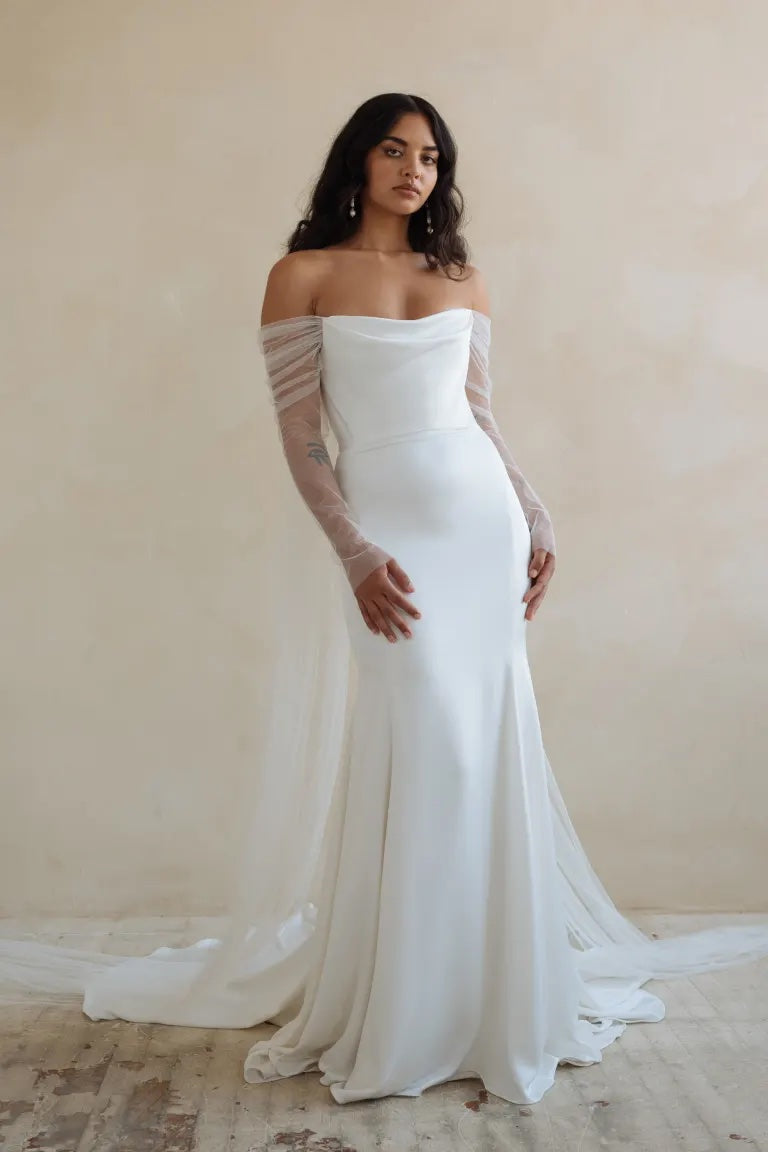 A woman in an elegant Jenny Yoo Wedding Dress featuring luxe satin fabric with off-the-shoulder sleeves stands against a neutral backdrop.