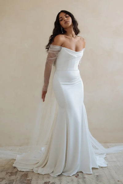 A woman posing in an elegant off-the-shoulder Olivia wedding dress made from luxe satin fabric with a flowing train by Bergamot Bridal.