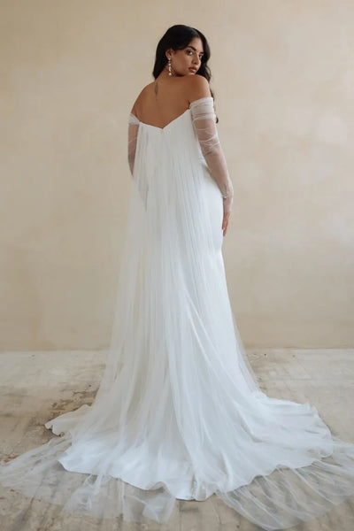 A woman in a luxe satin fabric Jenny Yoo Wedding Dress Olivia gown, an elegant white off-the-shoulder bridal dress with a fit and flare skirt, looking over her shoulder from Bergamot Bridal.