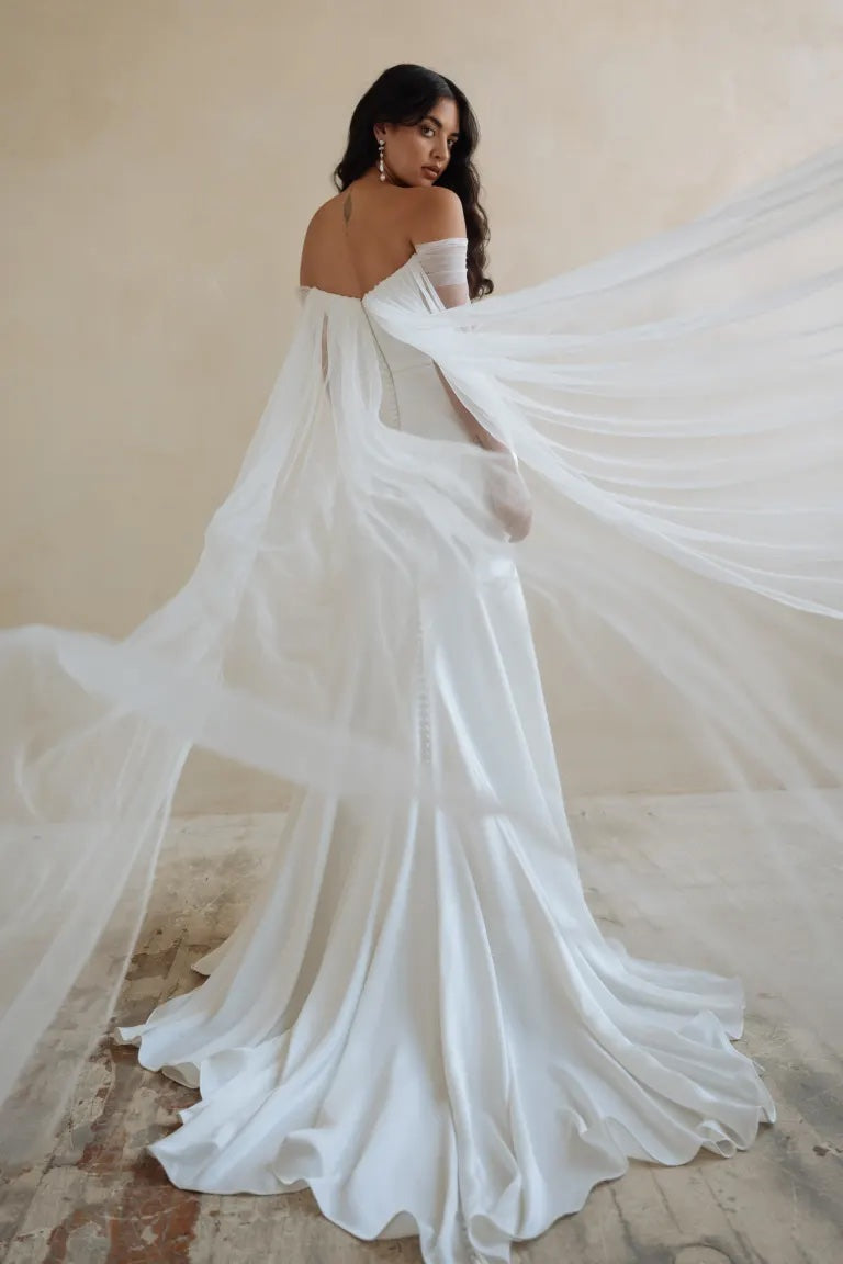 A bride in an elegant Olivia - Jenny Yoo Wedding Dress featuring a fit and flare skirt, made of luxe satin fabric, with a long flowing train and veil standing against a neutral backdrop from Bergamot Bridal.