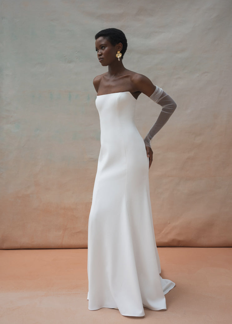 A person stands in a white, strapless bridal gown with Olivia Sleeves by Jenny Yoo from Bergamot Bridal, in front of a textured beige and grey backdrop.