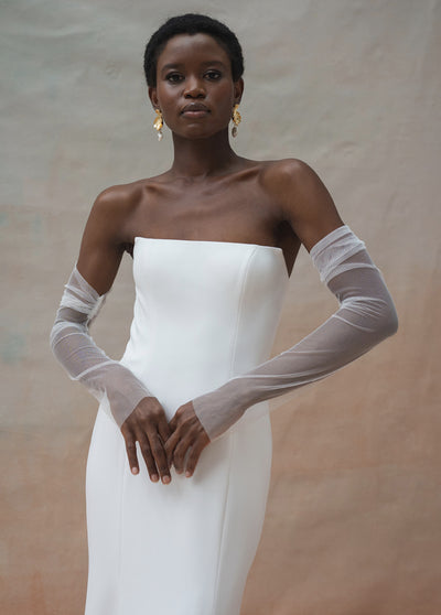 A woman wears Olivia Sleeves by Jenny Yoo - Off The Rack from Bergamot Bridal, styled with sheer mesh gloves, standing against a neutral background and looking at the camera.