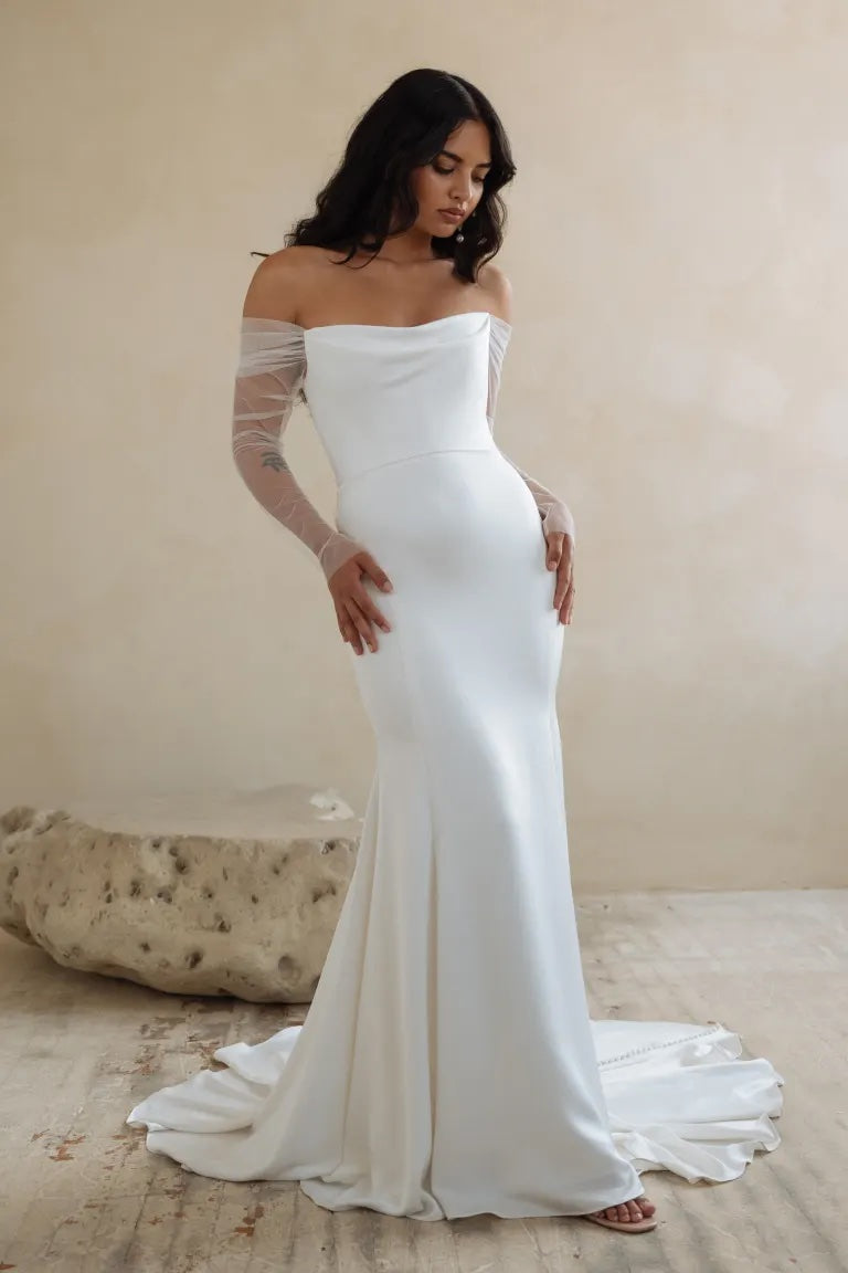 A person in an elegant off-the-shoulder Olivia - Jenny Yoo Wedding Dress gown made of luxe satin fabric with a fitted silhouette and a fit and flare skirt from Bergamot Bridal.