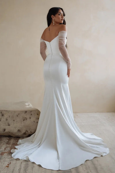 Woman in an elegant white off-the-shoulder Olivia - Jenny Yoo Wedding Dress gown made from luxe satin fabric, standing in a room with a neutral background.