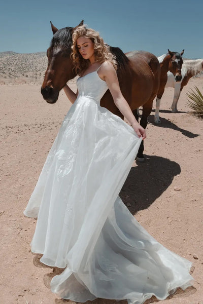 A woman in a romantic Abernathy - Jenny Yoo bridal gown standing beside horses in a desert setting.