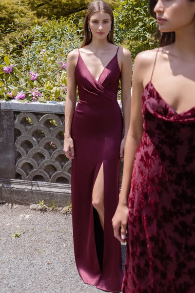 Two women in elegant burgundy satin Beckette bridesmaid dresses by Jenny Yoo standing outside, from Bergamot Bridal.