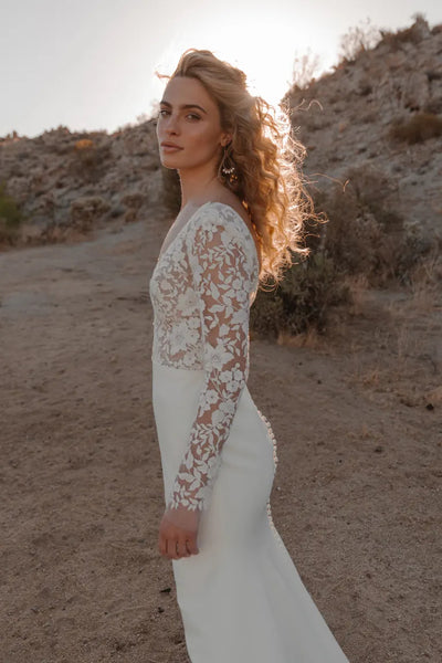 Woman in an elegant lace Julian - Jenny Yoo wedding dress with embroidered florals, standing on a desert path at sunset from Bergamot Bridal.