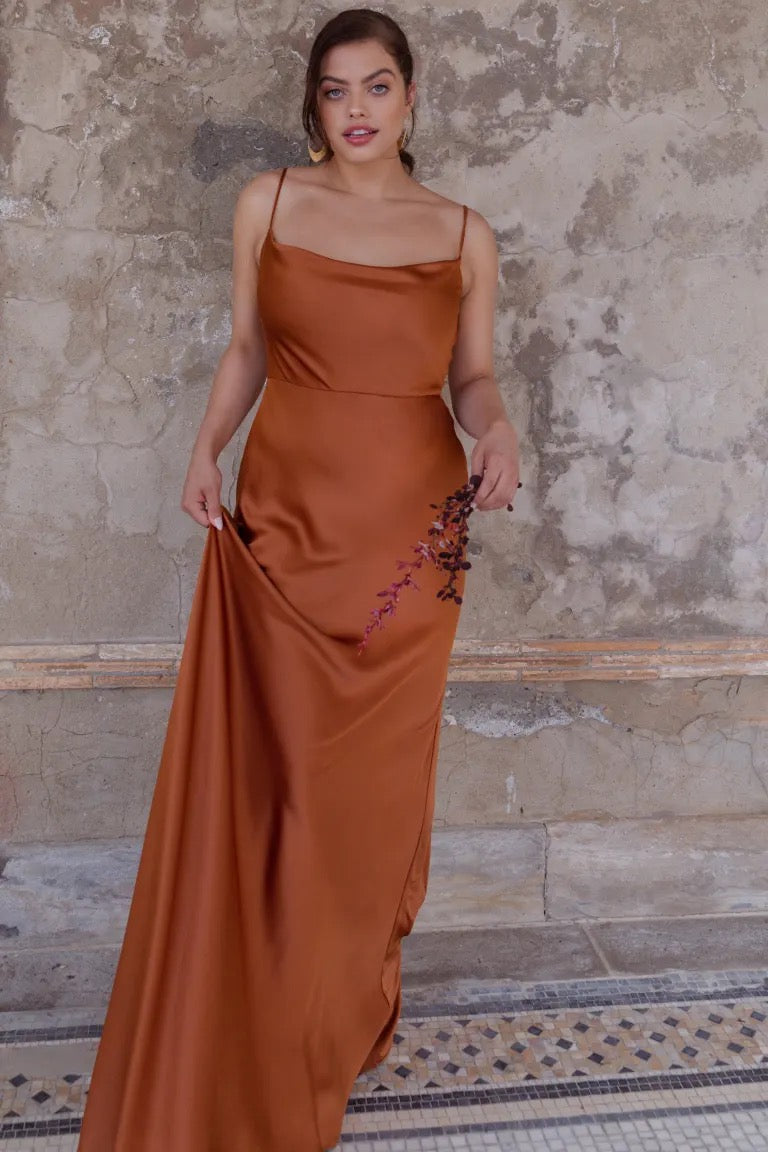 A woman in an elegant burnt orange Sylvie bridesmaid dress by Jenny Yoo, radiating Old Hollywood vibes, poses against a textured wall.