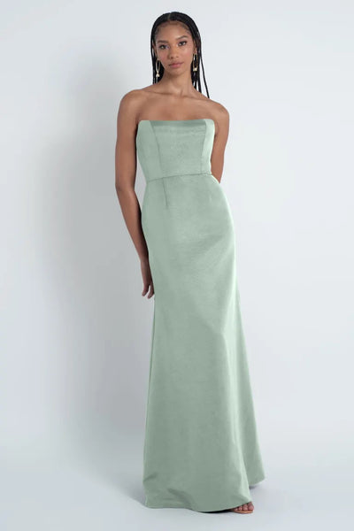 A woman in a strapless pastel green Paige - Jenny Yoo Bridesmaid Dress made of Luxe Faille fabric standing against a neutral background from Bergamot Bridal.