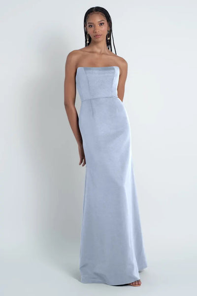 A woman wearing a strapless light blue Jenny Yoo Bridesmaid Dress made of Luxe Faille fabric stands against a neutral background from Bergamot Bridal.