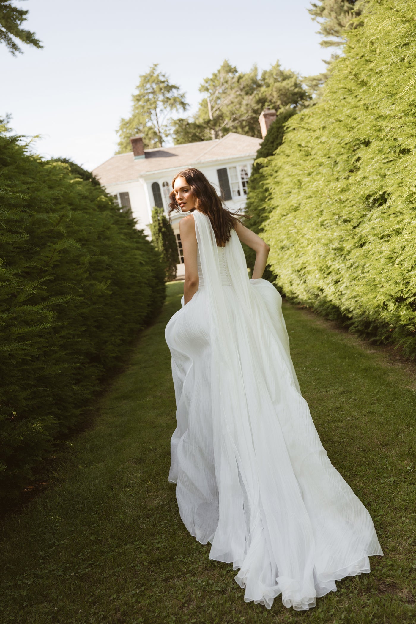 A woman in a long Penelope by Jenny Yoo gown from Bergamot Bridal walks along a grassy path between tall hedges, with a large house visible in the background.