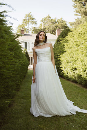 A woman in a strapless Penelope by Jenny Yoo gown from Bergamot Bridal stands on a grassy path between tall hedges, her iridescent chiffon dress softly shimmering, with a large house in the background.