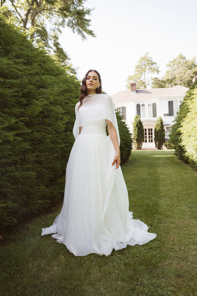 A woman in the iridescent chiffon Penelope by Jenny Yoo gown from Bergamot Bridal stands on a grassy path between tall green hedges, with a large white house in the background.