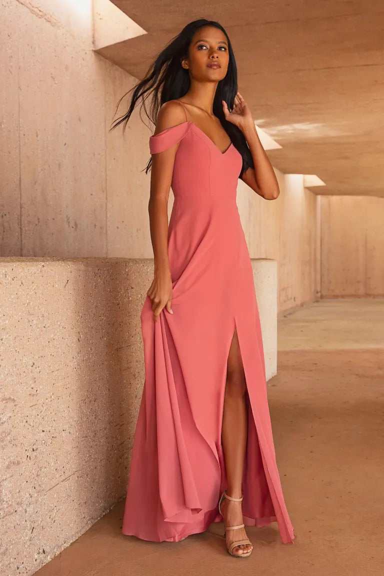 Woman in an elegant salmon pink chiffon Priya bridesmaid dress by Jenny Yoo with off-the-shoulder sleeves and a high slit, standing in a corridor with a warm-toned, textured wall from Bergamot Bridal.
