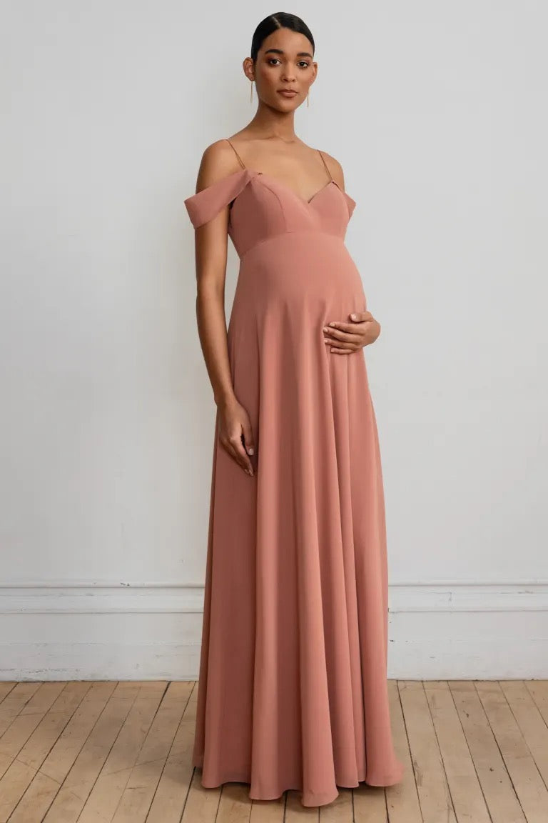 A pregnant woman posing in an elegant off-the-shoulder sleeve Priya maternity dress with a flowing A-line skirt by Jenny Yoo at Bergamot Bridal.