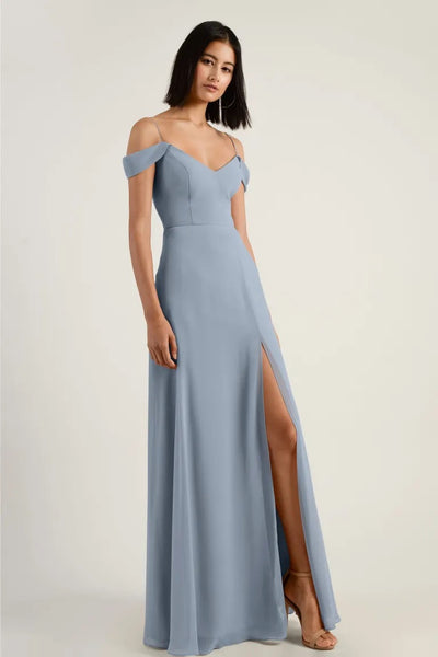 A woman in an elegant light blue chiffon bridesmaid dress with off-the-shoulder sleeves and a high side slit stands poised for a formal occasion wearing the Priya Bridesmaid Dress by Jenny Yoo from Bergamot Bridal.