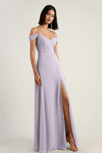 Woman posing in an elegant lilac chiffon bridesmaid dress with a high side slit and off-shoulder sleeves, the Priya Bridesmaid Dress by Jenny Yoo from Bergamot Bridal.