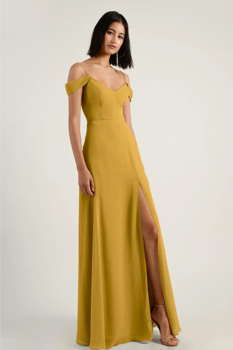 Woman posing in an elegant mustard yellow, off the shoulder sleeve Priya bridesmaid dress by Jenny Yoo from Bergamot Bridal with a thigh-high slit.