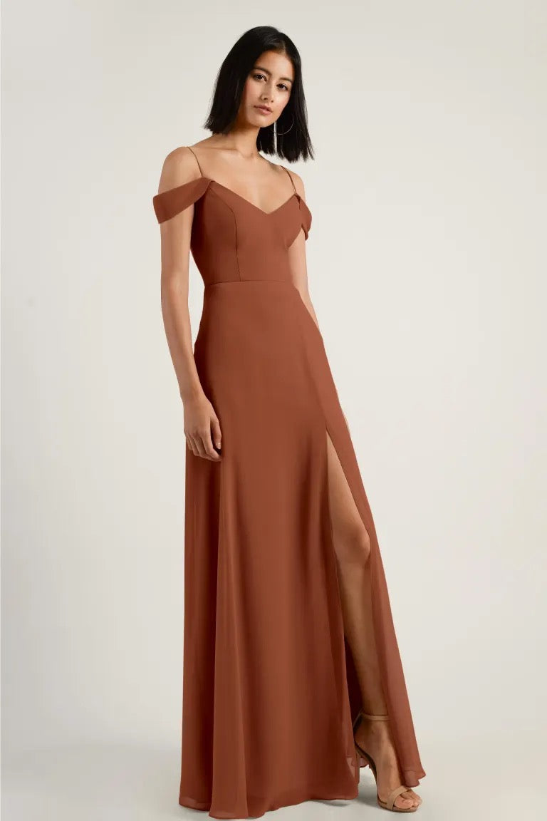 A woman wearing an elegant brown chiffon bridesmaid dress with off-the-shoulder sleeves and a high thigh slit, known as the Priya Bridesmaid Dress by Jenny Yoo from Bergamot Bridal.