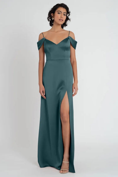 A woman in a Priyanka bridesmaid dress by Jenny Yoo in an elegant green satin with an off-the-shoulder sleeve and thigh-high slit stands poised against a plain background.