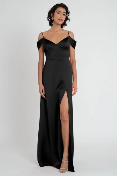 A woman in a Priyanka - Bridesmaid Dress by Jenny Yoo in a black satin evening gown with a side slit and off-the-shoulder straps stands against a plain background for an elegant look from Bergamot Bridal.