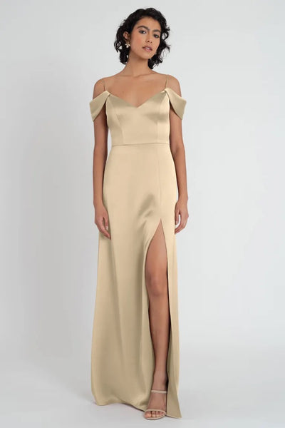 A woman wearing an elegant beige satin evening gown with off-the-shoulder sleeves and a high leg slit, the Priyanka bridesmaid dress by Jenny Yoo from Bergamot Bridal.