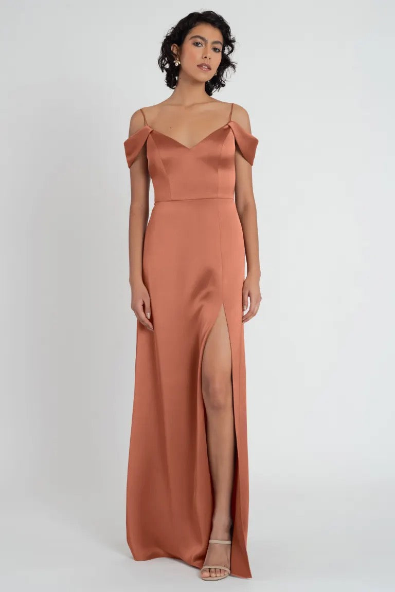 A woman posing in an elegant satin evening gown with off-the-shoulder sleeves and a side slit, the Priyanka Bridesmaid Dress by Jenny Yoo from Bergamot Bridal.