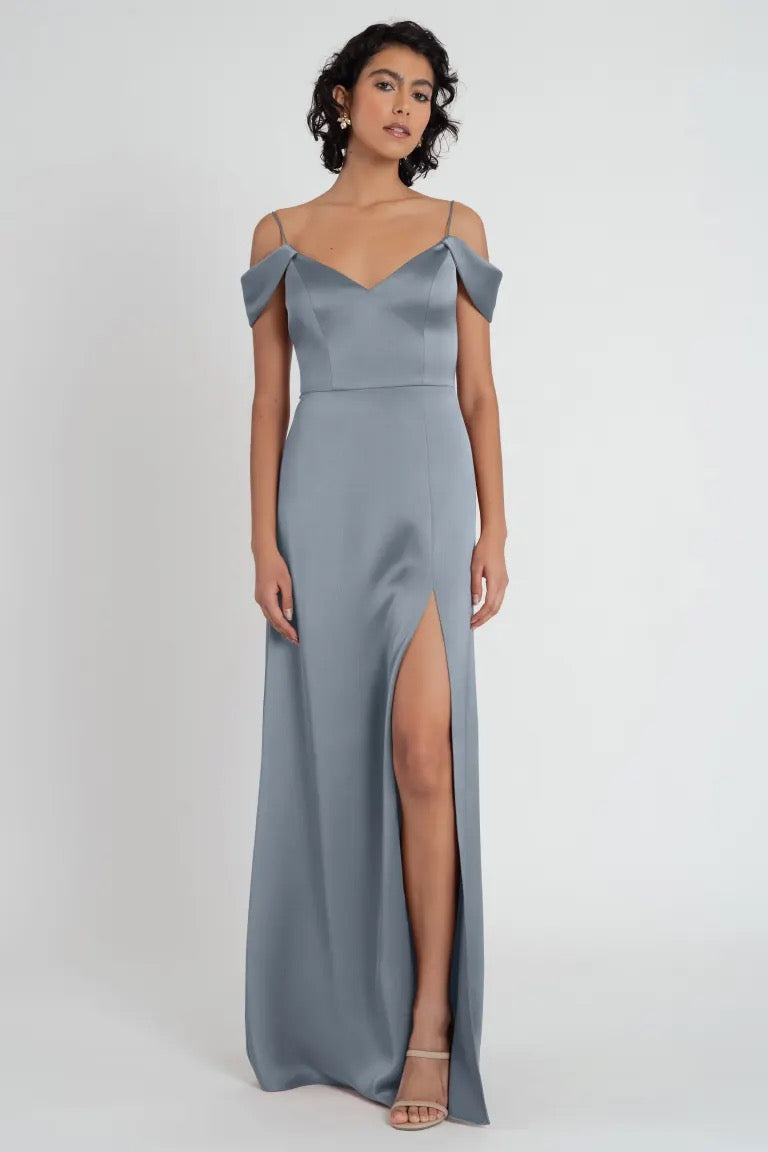 Woman posing in an elegant gray satin Priyanka bridesmaid dress by Jenny Yoo with off-the-shoulder sleeves and a side slit from Bergamot Bridal.