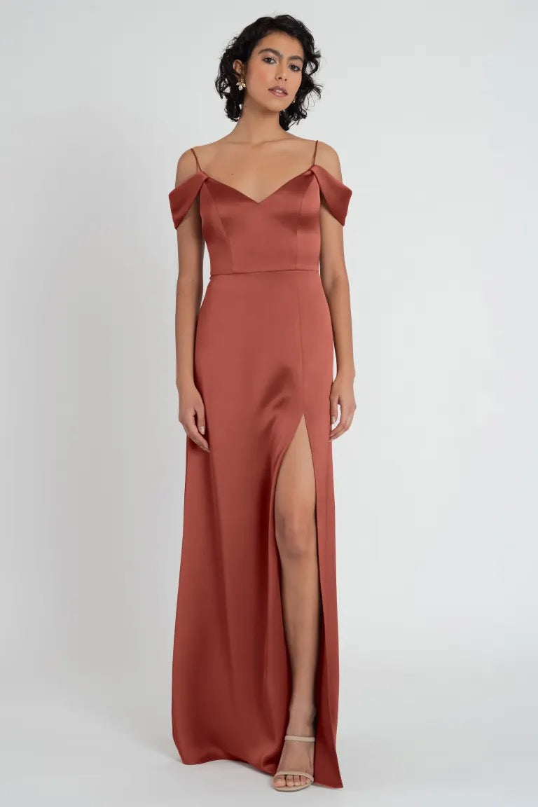 A woman stands posing in a formal rust-colored satin bridesmaid dress with off-the-shoulder sleeves and a side slit, the Priyanka Bridesmaid Dress by Jenny Yoo from Bergamot Bridal.