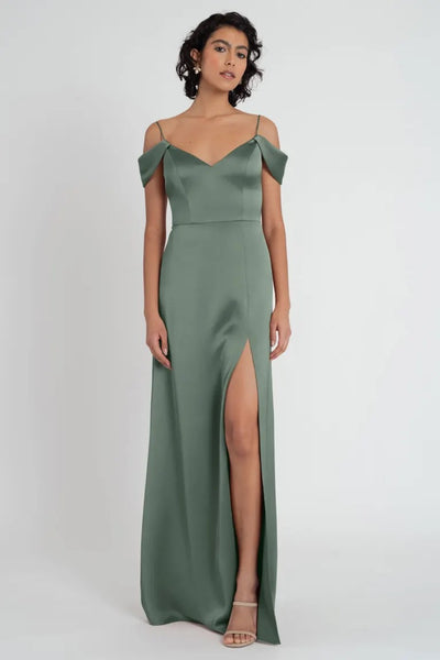 A woman in an elegant green satin Priyanka bridesmaid dress by Jenny Yoo with a thigh-high side slit poses against a neutral background from Bergamot Bridal.