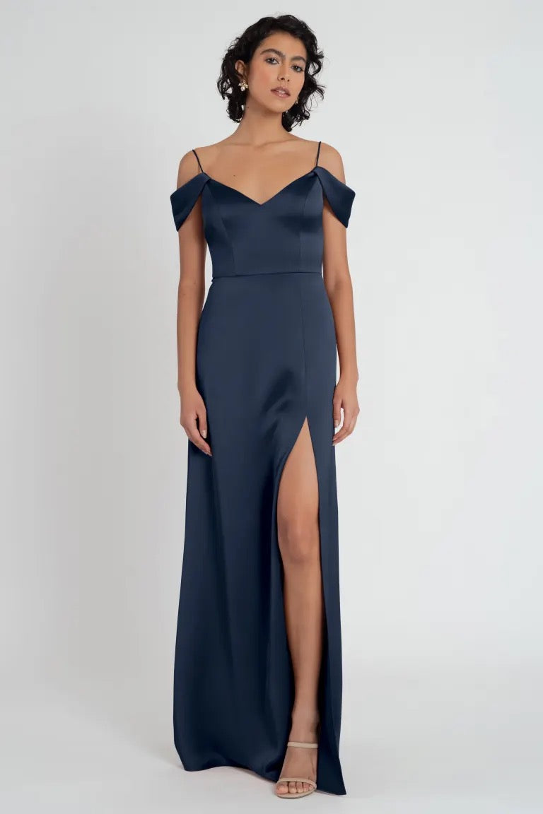 A woman in an elegant navy blue off the shoulder evening gown with a side slit, the Priyanka Bridesmaid Dress by Jenny Yoo from Bergamot Bridal.