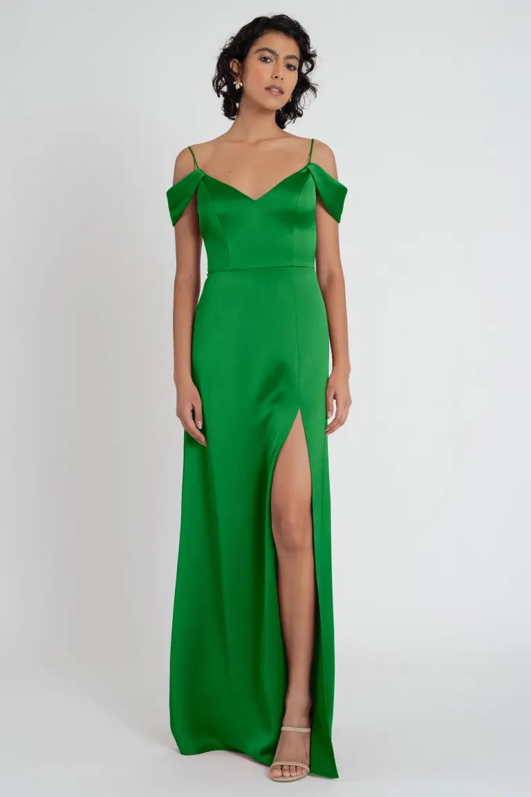 A woman in a Priyanka - Bridesmaid Dress by Jenny Yoo with a thigh-high slit and off-the-shoulder sleeves stands against a white background from Bergamot Bridal.