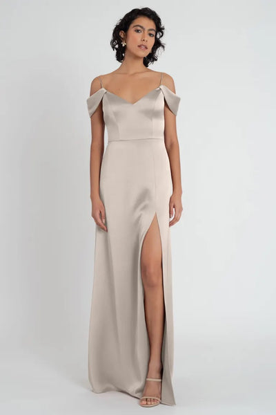 A model wearing an elegant beige satin Priyanka bridesmaid dress by Jenny Yoo with a side slit and off-shoulder straps from Bergamot Bridal.