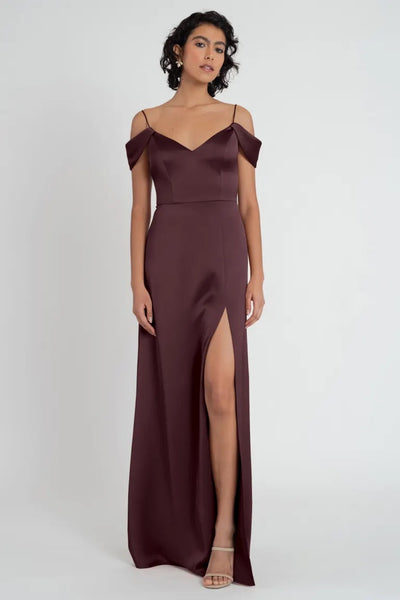 Woman posing in an elegant off-the-shoulder Priyanka - Bridesmaid Dress by Jenny Yoo evening gown in burgundy with a side slit from Bergamot Bridal.