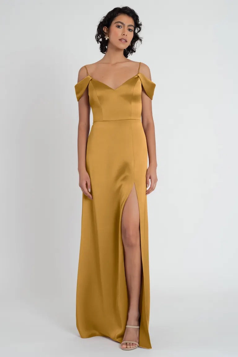 Woman posing in an elegant mustard satin V-neck Priyanka - Bridesmaid Dress by Jenny Yoo evening gown with off-the-shoulder sleeves and a side slit from Bergamot Bridal.