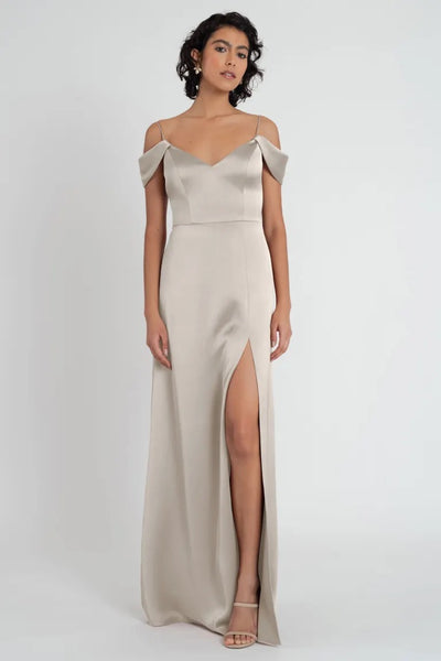 A woman in a chic beige satin V-neck bridesmaid dress with off-the-shoulder sleeves and a thigh-high slit, the Priyanka Bridesmaid Dress by Jenny Yoo from Bergamot Bridal.